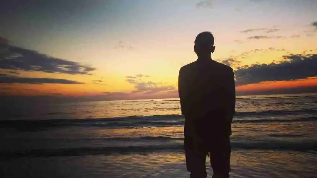 Yasin Haider Sami, seen from behind in silhouette, standing at the edge of the sea at sunset.